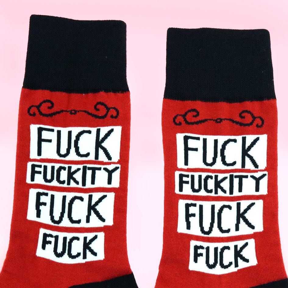 A pair of socks standing against a pink background. The socks are red and black and read Fuck Fuckity Fuck Fuck.