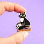 A hard enamel lapel pin being held in a hand. The pin is in the shape of a black and white cat with a scull, sitting on books. The pin reads Cat’s and Books.