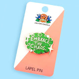 A hard enamel pin on Jubly-Umph card stock with a blue background. The pin is green glitter with white stars and reads Embrace the Chaos.