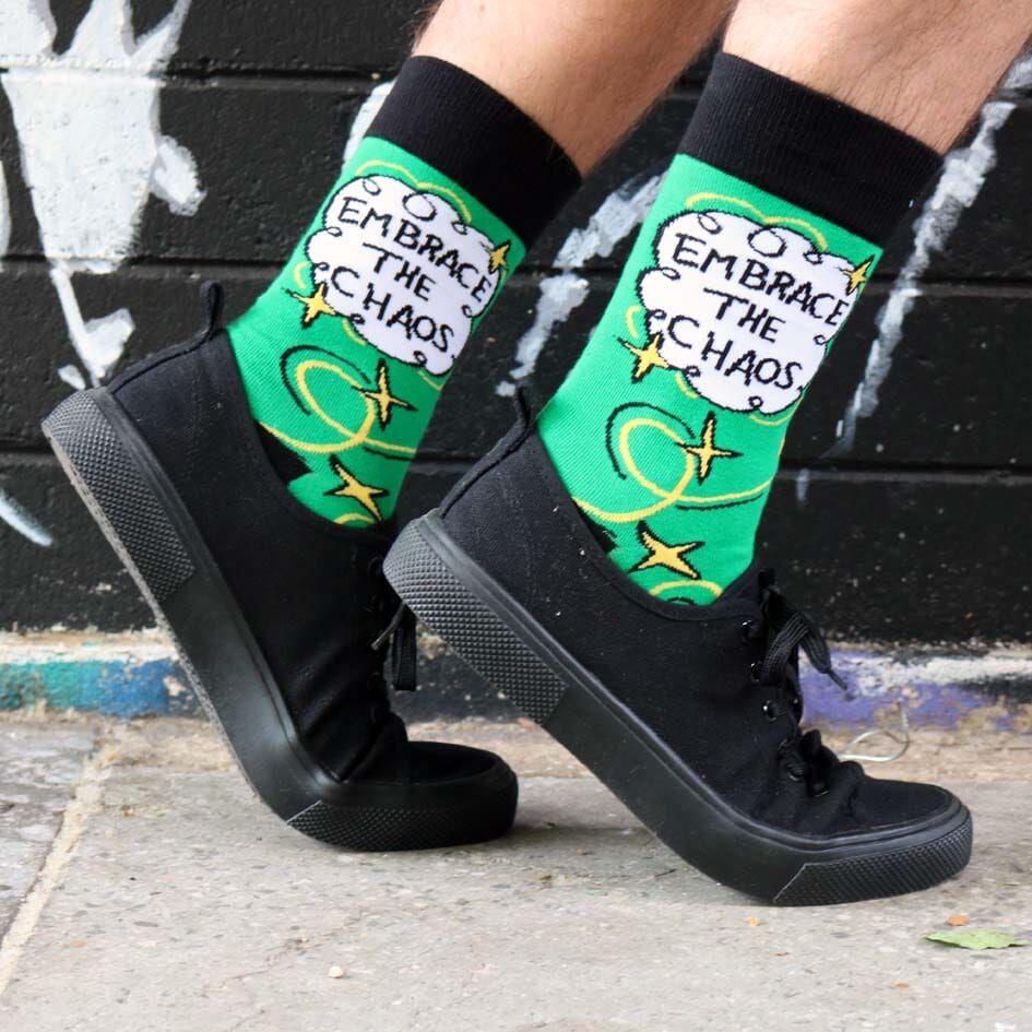 A pair of socks being worn with black shoes standing against a graffiti wall. The socks are green and gold and read Embrace The Chaos. 