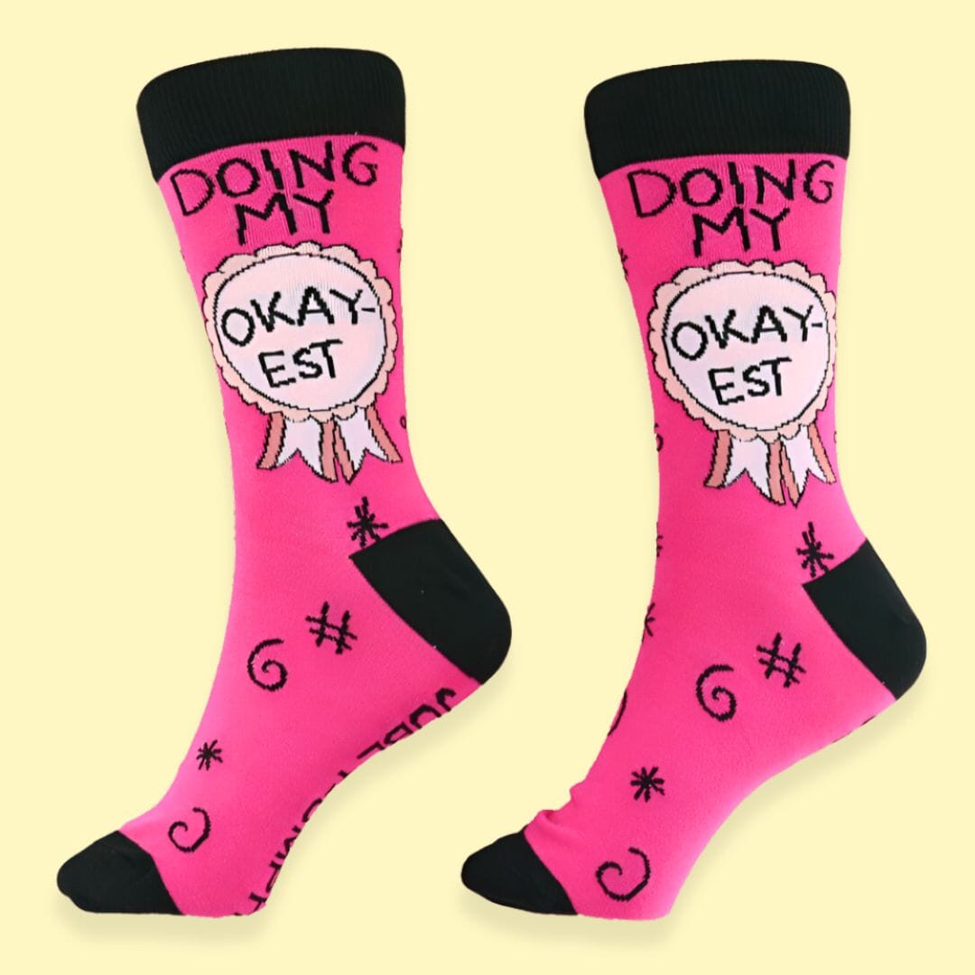 A pair of socks standing against a yellow background. The socks are pink and black and read Doing My Okay-est.