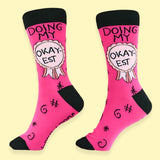 A pair of socks standing against a yellow background. The socks are pink and black and read Doing My Okay-est.