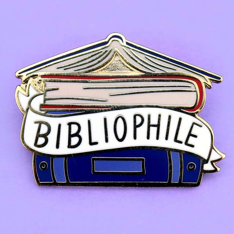 A hard enamel lapel pin on a purple background. The pin says Bibliophile in the middle of a stack of blue books.