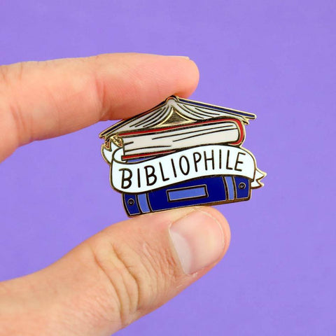 A hard enamel lapel pin being held in a hand against a purple background. The pin says Bibliophile in the middle of a stack of blue books.