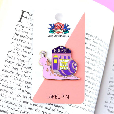 A hard enamel lapel pin on Jubly-Umph cardstock. The pin says say Bookish. The pin design is a snail with a bookshop on its back.