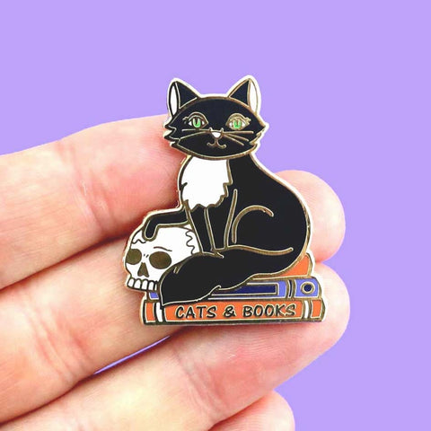 A hard enamel lapel pin being held in a hand. The pin is in the shape of a black and white cat with a scull, sitting on books. The pin reads Cat’s and Books.