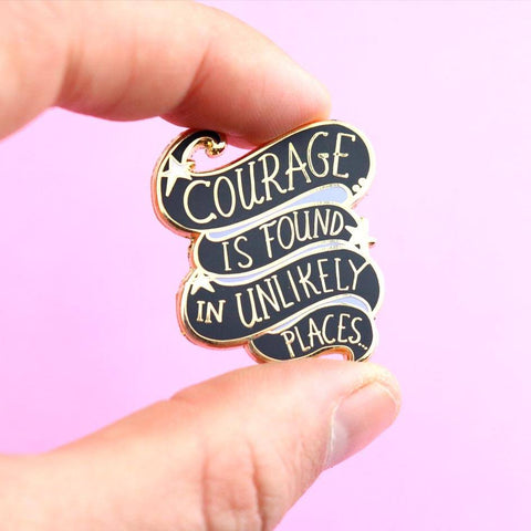 A hard enamel lapel pin being held in a hand. The pin is black and purple and reads Courage Is Found In Unlikely Places.