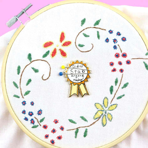 A hard enamel magnetic needle minder displayed on an embroidery hoop with two sewing needles. The pin is in the shape of an award ribbon. The ribbon is yellow and white, and reads Didn’t Stab Anyone Today!