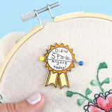 A hard enamel magnetic needle minder displayed on an embroidery hoop with two sewing needles. The pin is in the shape of an award ribbon. The ribbon is yellow and white, and reads Didn’t Stab Anyone Today!