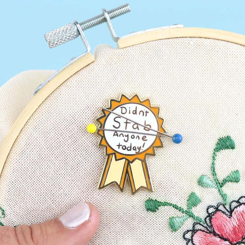 A hard enamel magnetic needle minder displayed on an embroidery hoop with two sewing needles. The pin is in the shape of an award ribbon. The ribbon is yellow and white, and reads Didn’t Stab Anyone Today!