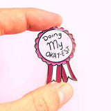A hard enamel lapel pin being held in a hand. The pin is in the shape of an award ribbon. The ribbon is dark pink and light pink and reads Doing my Okay-est.