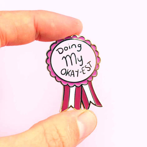 A hard enamel lapel pin being held in a hand. The pin is in the shape of an award ribbon. The ribbon is dark pink and light pink and reads Doing my Okay-est.