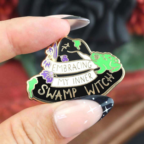 A hard enamel pin being held in hand. The pin is black in the shape of a witches hat. The hat reads Embracing My Inner Swamp Witch.