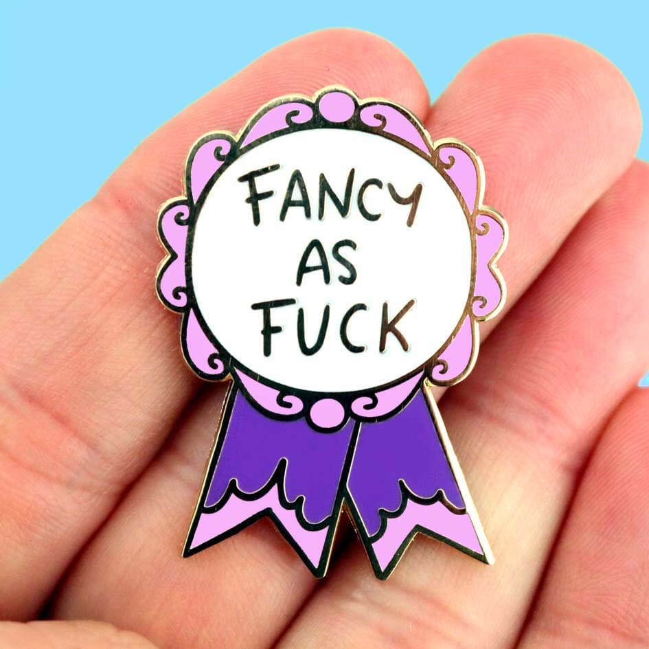 A hard enamel lapel pin being held in a hand. The pin is in the shape of a pink and purple ribbon award and reads Fancy As Fuck.