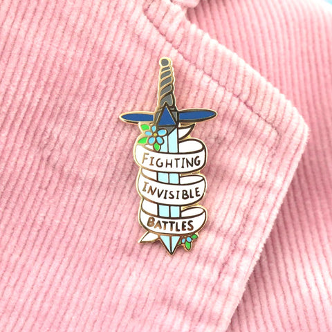 A hard enamel pin on a pink jacket. The pin is in the shape of a dagger and reads Fighting Invisible Battles.