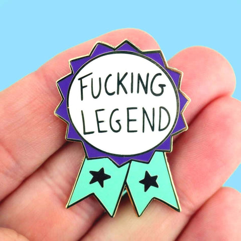 A hard enamel lapel pin being held in a hand. The pin is in the shape of a teal and purple ribbon award and reads Fucking Legend.