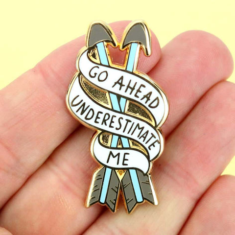 A hard enamel lapel pin being held in a hand. The pin is in the shape of two arrows and reads Go Ahead Underestimate Me.