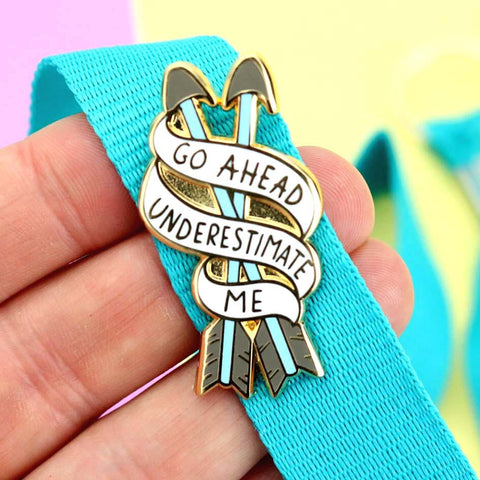 A hard enamel lapel pin being held in a hand on a blue lanyard. The pin is in the shape of two arrows and reads Go Ahead Underestimate Me.