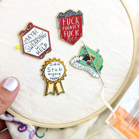 A group of four hard enamel magnetic needle minders displayed on an embroidery hoop with two sewing needles. The first pin is in the shape of an award ribbon. The ribbon is yellow and white, and reads Didn’t Stab Anyone Today! The other three needle minders read Maybe Swearing Will Help, Fuck Fuckity Fuck Fuck and Sewciopath.