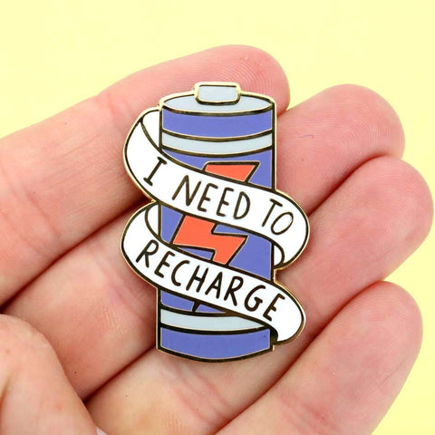 A hard enamel lapel pin held in a hand. The lapel pin is in the shape of a battery and reads I Need To Recharge.