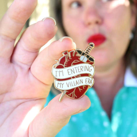 A hard enamel lapel pin being held in a hand. The lapel pin is in the shape of a human heart with a dagger. The pin reads I'm Entering My Villain Era.