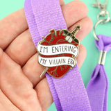 A hard enamel lapel displayed on a purple lanyard. The lapel pin is in the shape of a human heart with a dagger. The pin reads I'm Entering My Villain Era.