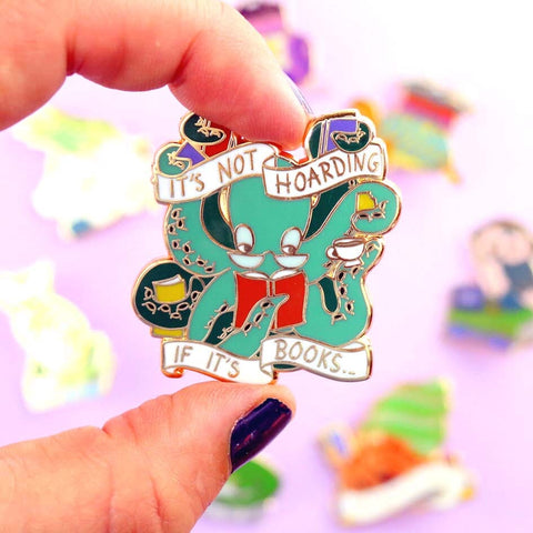 A hard enamel lapel pin being held in a hand. The pin is in the shape of an Octopus carrying books. The pin reads It's Not Hoarding If It's Books.