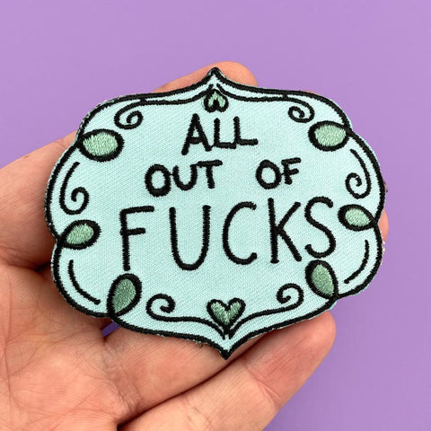 An iron on embroidered patch  being held in a hand against a purple background. The patch is blue and green and reads All Out Of Fucks.