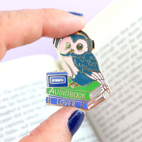 A hard enamel lapel pin being held in a hand. The pin is in the shape of an owl sitting on a stack of books wearing headphones. The pin reads Audiobook Lover.