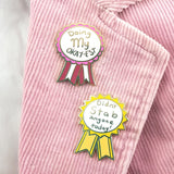 Two hard enamel lapel pins on a pink jacket. The first pin is in the shape of an award ribbon. The ribbon is dark pink and light pink and reads Doing my Okay-est. The second pin is in the shape of an award ribbon and is yellow and white and reads Didn’t Stab Anyone Today!