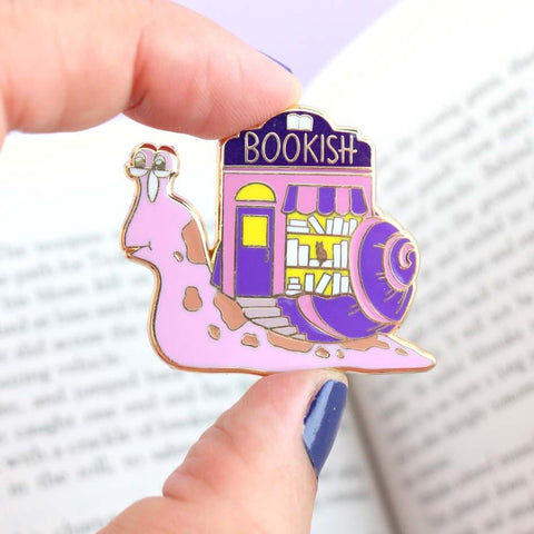 A hard enamel lapel pin being held in a hand. The pin says say Bookish. The pin design is a snail with a bookshop on its back.