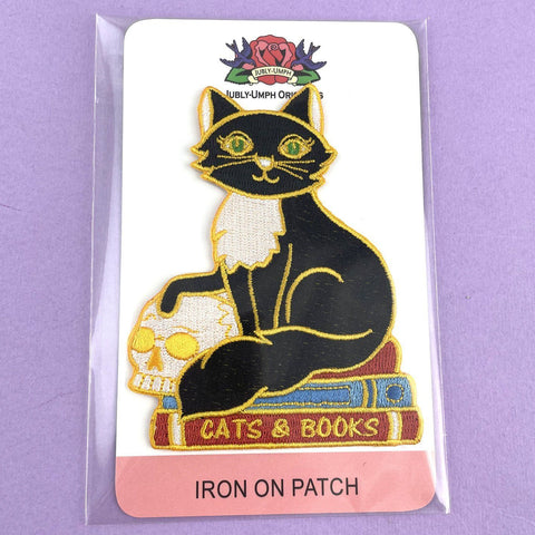 An iron on embroidered patch on Jubly-Umph cardstock against a purple background. The patch is a black and white cat with a scull sitting on books. The patch reads Cat’s and Books.