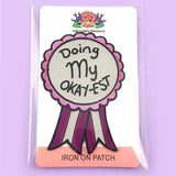 An iron on embroidered patch on Jubly-Umph cardstock. The patch is in the shape of an award ribbon. The ribbon is pink and grey and reads Doing my Okay-est.