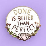 A hard enamel lapel pin on a light purple background. The pin is white with gold lettering and reads Done Is Better Thank Perfect.