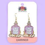 A pair of dangle earrings displayed on on Jubly-Umph cardstock. Displayed on purple background. The earrings are purple and in the shape of a bottle with clouds and lightning bolts. The earrings read Do No Harm Take No Shit.