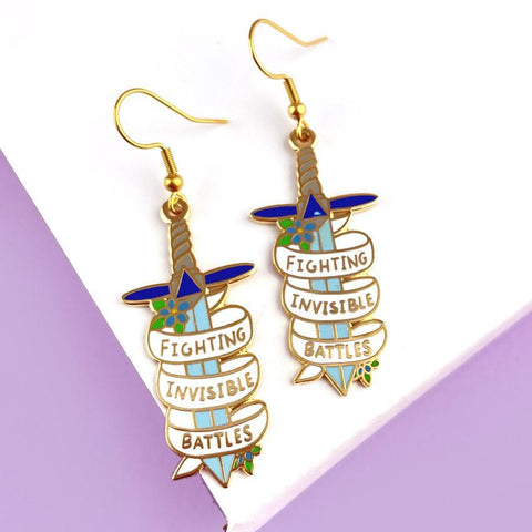 A pair of earrings displayed on a purple background. The earrings are in the shape of dagger and read Fighting Invisible Battles.