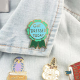 A hard enamel lapel pin displayed on a denim jacket with other Jubly-Umph lapel pins. The pin is in the shape of a green award ribbon. The pin reads Got Dressed Today.